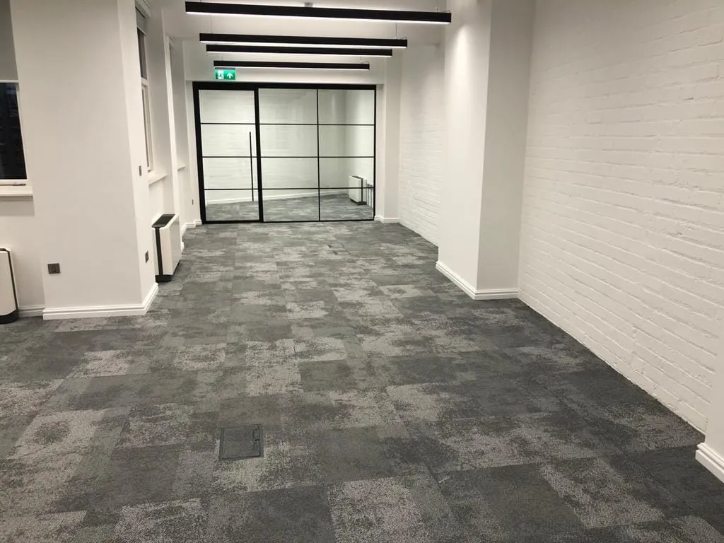 Empty office space with gray patterned carpet tiles, white brick walls, and a glass door at the far end. Overhead fluorescent lights illuminate the room.