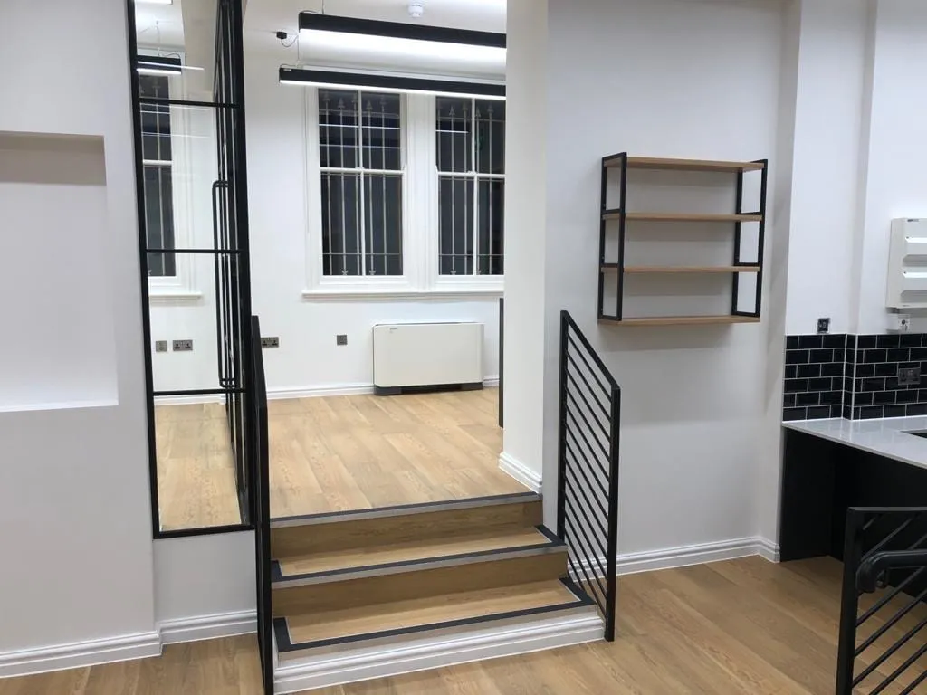 Modern interior with wooden floors, a black metal-framed mirror, stairs leading to a raised area, and wall-mounted shelves. Sleek and minimalist design.
