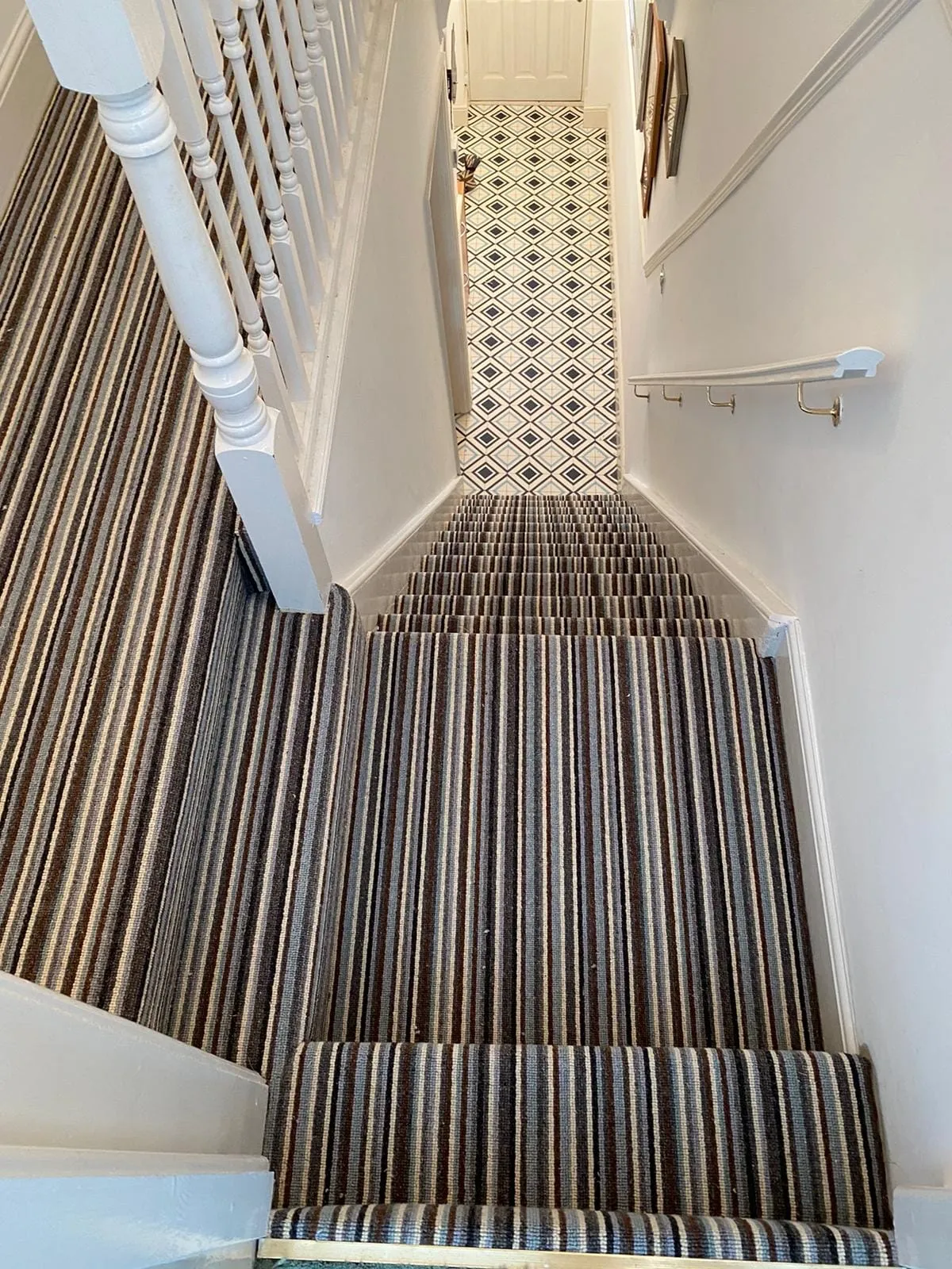 Staircase with brown and beige striped carpet leads to a hallway with black and white diamond-patterned tiles. White walls and handrails line the stairs.