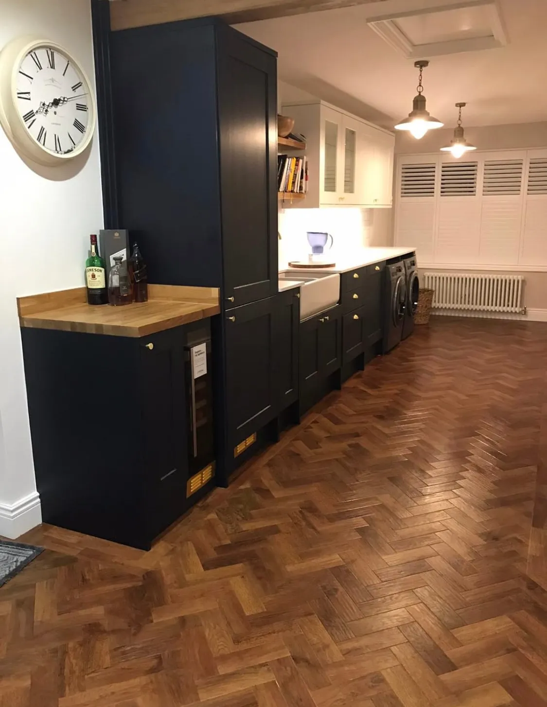 Sleek kitchen with dark cabinets, wooden herringbone karndean floor, and a white farmhouse sink. Vintage clock and pendant lights add rustic charm.