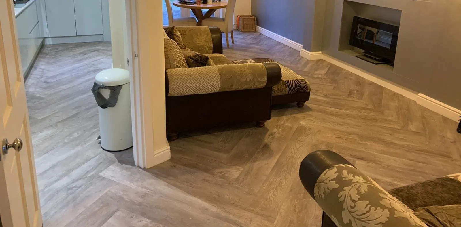 Spacious living room with wood-patterned karndean flooring, cozy patterned sofas, a fireplace, and a view into the kitchen and dining area with soft lighting.