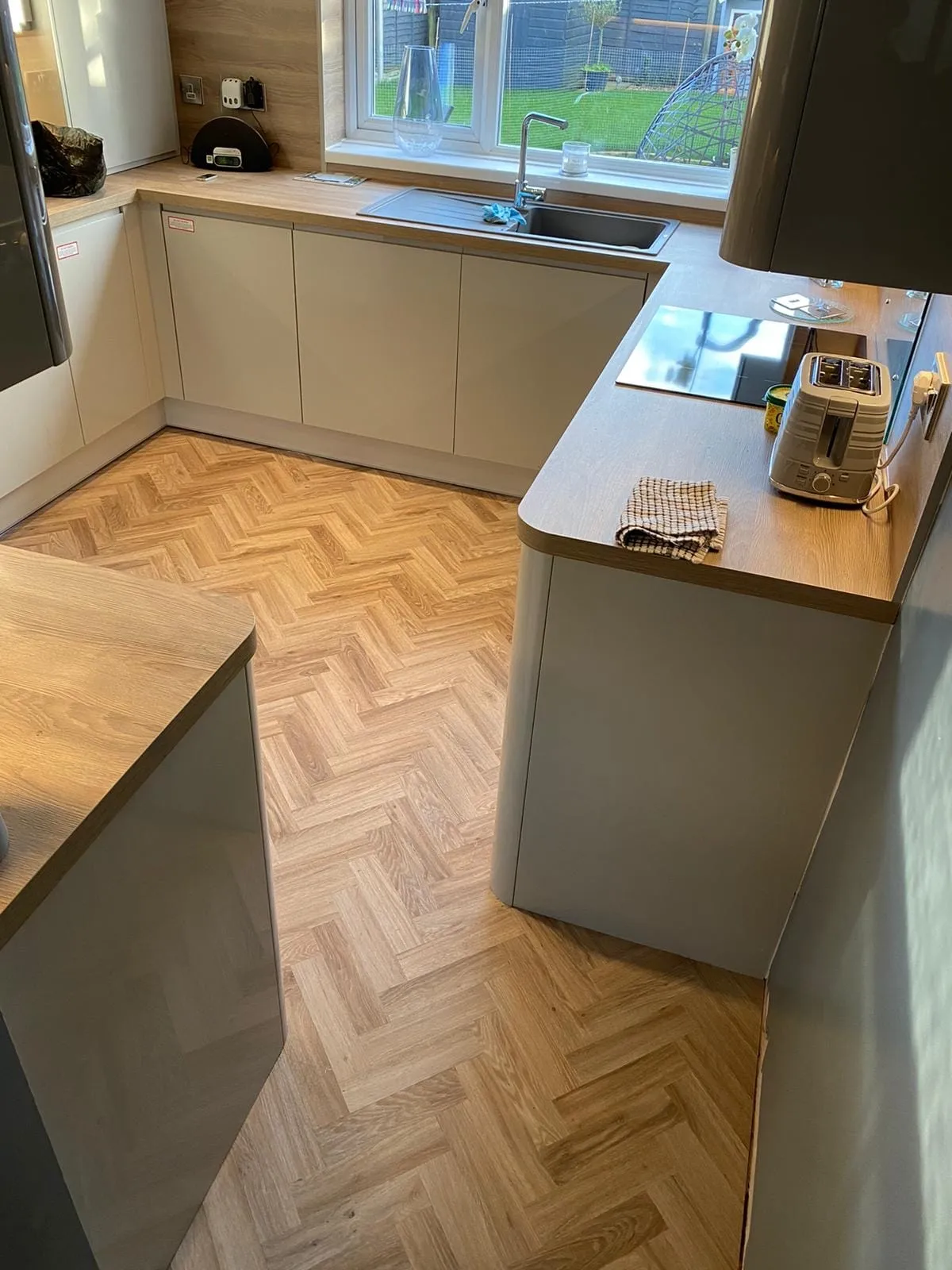 Modern kitchen with light wood LVT floor, gray cabinets, and wooden countertops. Toaster and dish towel on counter near stovetop, sink by window.