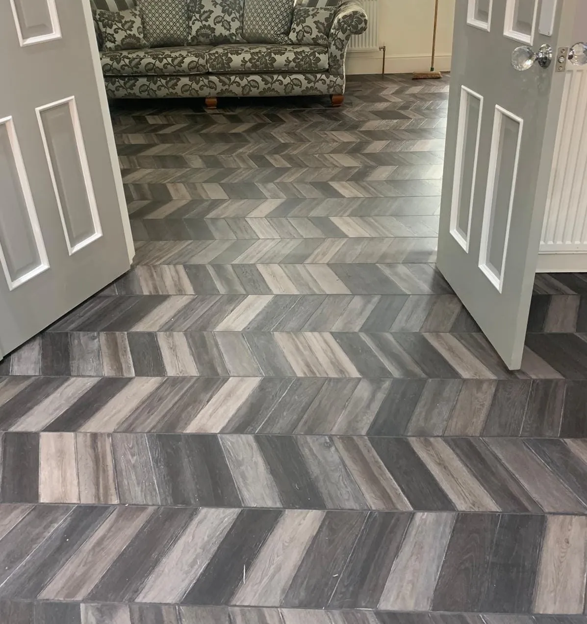 Herringbone-patterned LVT flooring Solihull that leads to an open-door room with a floral-patterned sofa, creating a warm and inviting ambiance.