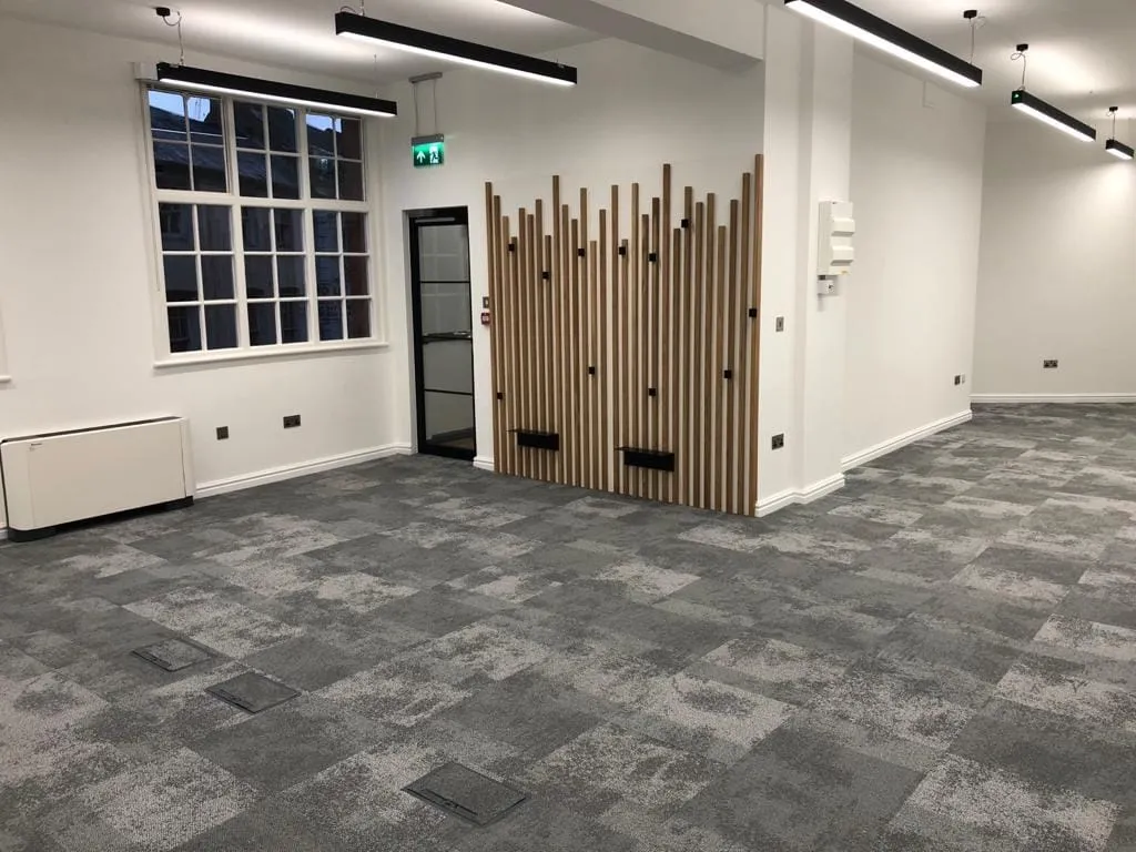 Modern office space with gray carpet tiles and white walls. A wooden slat wall feature adds texture. Large windows provide natural light.