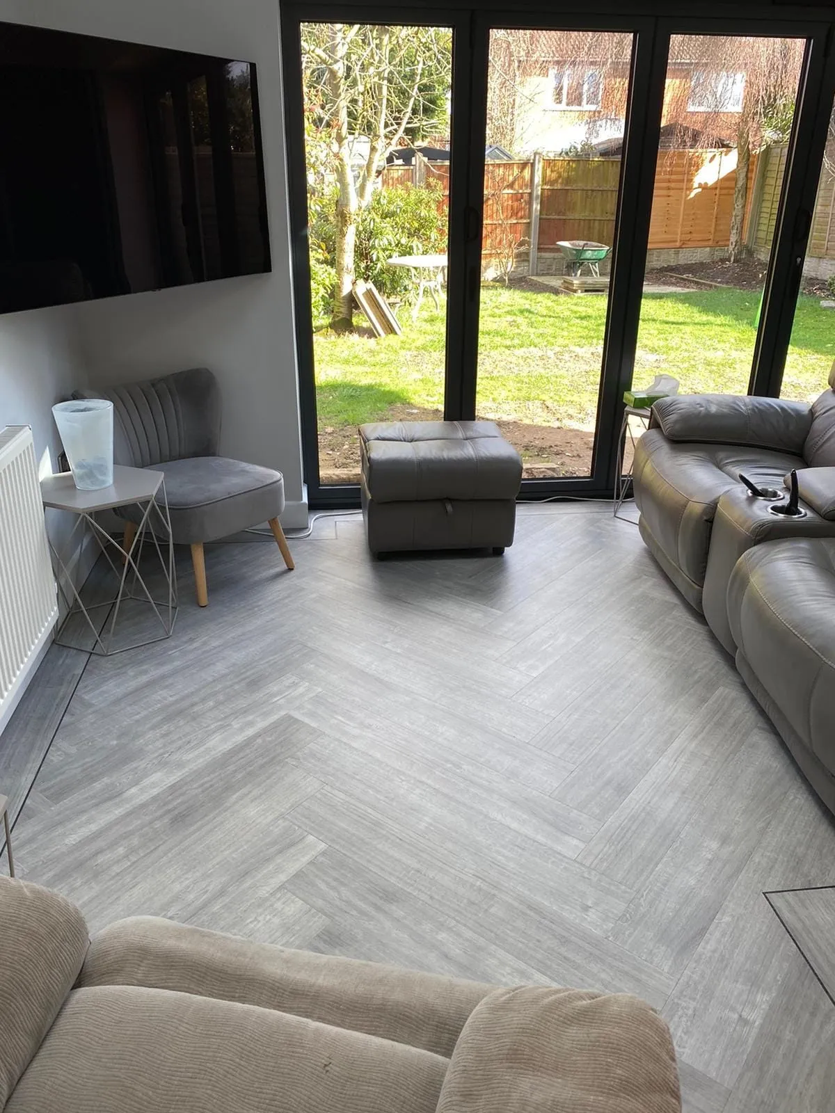 A cozy living room with gray herringbone flooring. Large windows overlook a sunny garden. The room has gray seating, a wall-mounted TV, and light decor.